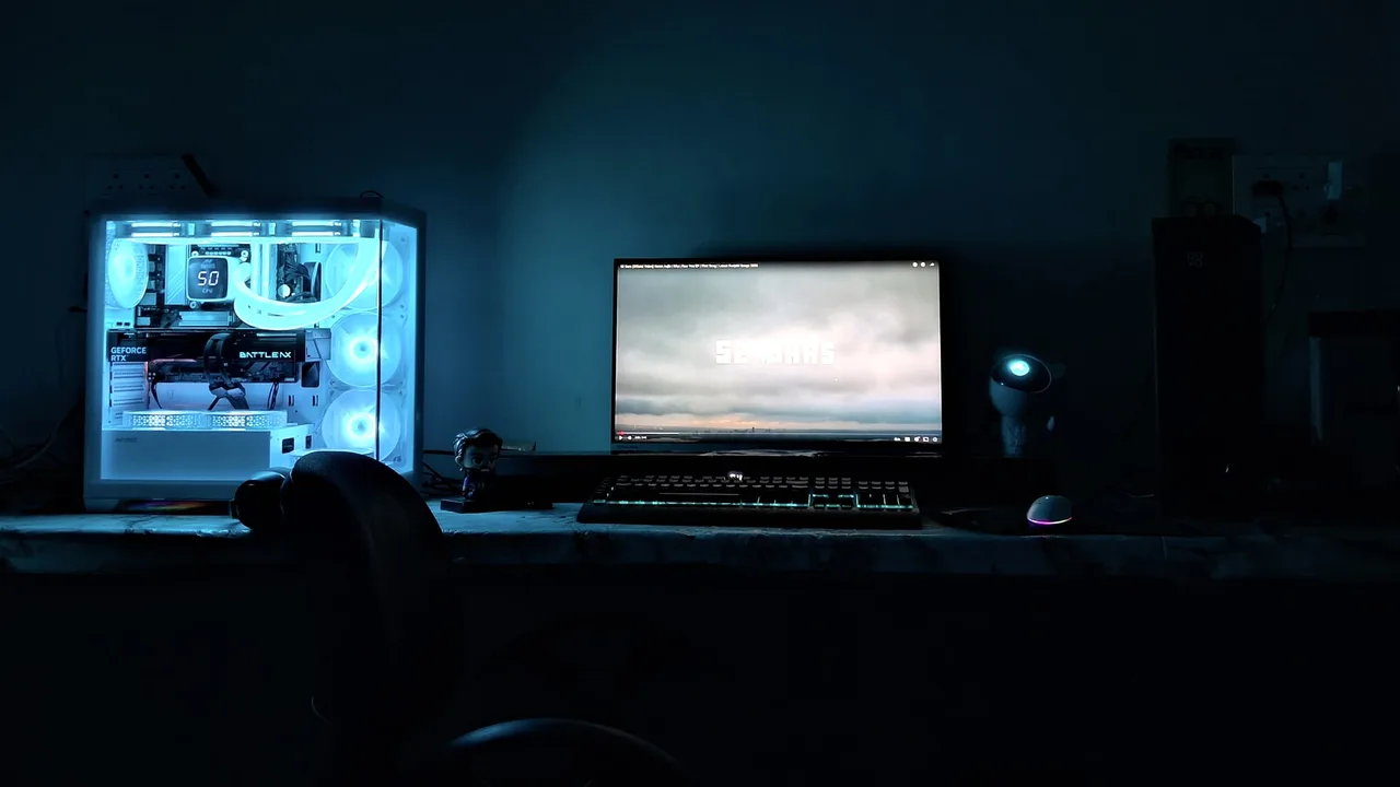 A dim desk setup with a white glass-panel PC case lit in blue on the left, showing a GeForce RTX graphics card and liquid-cooling tubes, next to a monitor, an RGB mechanical keyboard and a glowing mouse